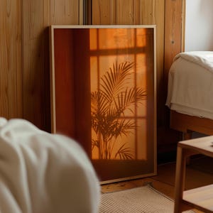 May include: A rectangular framed wall art piece featuring a warm, amber-toned photograph of a palm plant silhouette cast against a window pane. The print is encased in a light-colored wooden frame, displaying deep orange and brown hues.