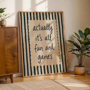 May include: A rectangular framed wall art piece featuring a cream-colored center panel with a scalloped edge, surrounded by dark blue vertical stripes. The text "actually it's all fun and games" is printed in dark blue, handwritten-style lettering.