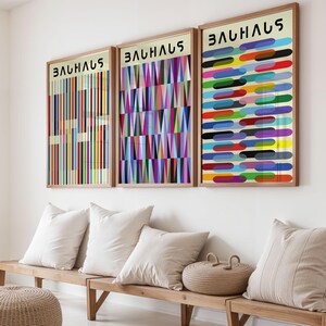 Puede incluir: Tres láminas enmarcadas de arte Bauhaus con diseños geométricos coloridos cuelgan sobre un banco de madera con cojines blancos. Las láminas presentan líneas verticales, triángulos y formas horizontales en varios colores. La palabra "BAUHAUS" está impresa en la parte superior de cada marco.