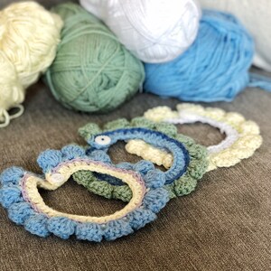 May include: Four crocheted headbands in different colors, including blue, green, yellow, and white. The headbands are arranged in a semi-circle on a gray surface.