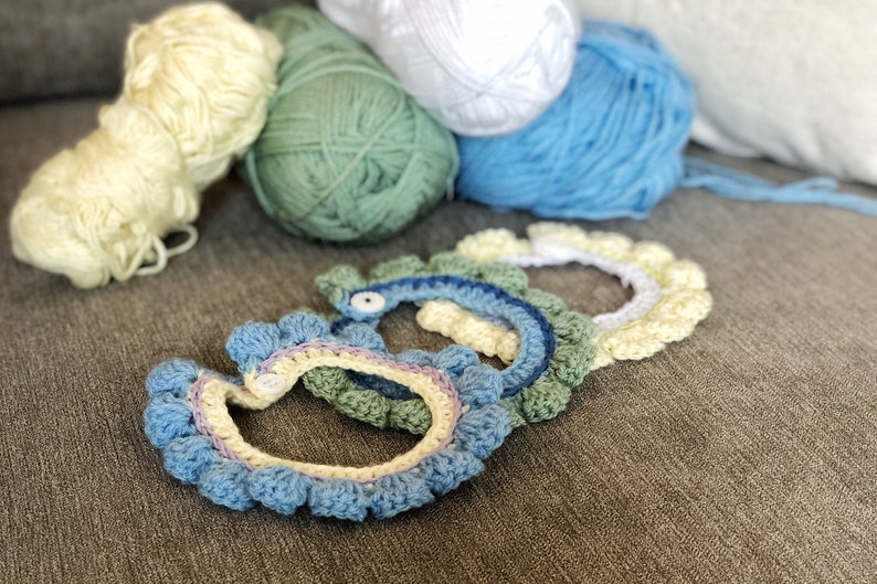 May include: Four crocheted headbands in different colors, including blue, green, yellow, and white. The headbands are arranged in a semi-circle on a gray surface.