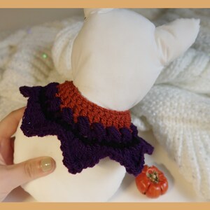May include: A purple and orange crocheted collar for a pet. The collar is in the shape of a star with a ruffled edge.