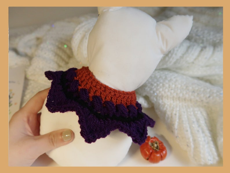 May include: A purple and orange crocheted collar for a pet. The collar is in the shape of a star with a ruffled edge.
