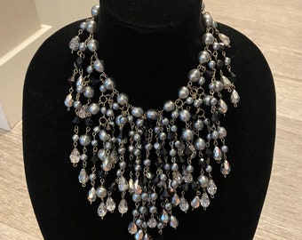 May include: A silver-toned statement necklace with cascading strands of faux pearls and crystal-like beads. The necklace is displayed on a black velvet bust, showcasing its intricate design and elegant draping.
