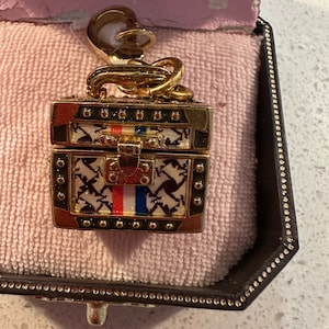 Puede incluir: Un charm de color dorado en forma de una pequeña maleta vintage adornada con un diseño de patrón blanco y negro. La maleta tiene una franja roja, blanca y azul en la parte delantera.