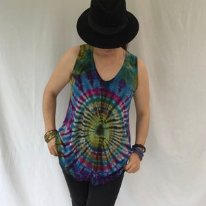Op de afbeelding: Een tie-dye tanktop met een cirkelvormig patroon in tinten blauw, groen, paars en roze. Het tanktop heeft een v-hals en is mouwloos. Het model draagt een zwarte broek en een zwarte fedora.