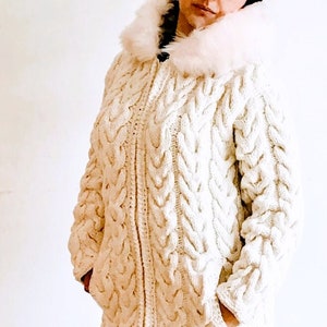 Puede incluir: Un cárdigan blanco de punto de cable con capucha de piel sintética. El cárdigan tiene cierre de cremallera y dos bolsillos.