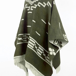 May include: Olive green poncho with white geometric patterns and fringe trim. The poncho features a repeating design of rectangles, lines, and floral shapes. The edges are finished with a white fringe.