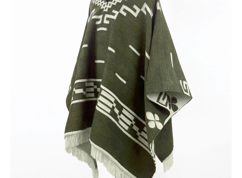May include: Olive green poncho with white geometric patterns and fringe trim. The poncho features a repeating design of rectangles, lines, and floral shapes. The edges are finished with a white fringe.