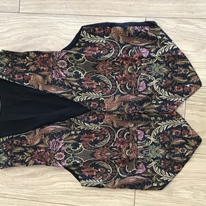 K&ouml;nnte beinhalten: Eine &auml;rmellose Weste mit dunkelblauer Grundfarbe und einem komplexen, mehrfarbigen Brokatmuster aus Blumen und V&ouml;geln in Rot-, Gold-, Gr&uuml;n- und Violettt&ouml;nen. Das Kleidungsst&uuml;ck verf&uuml;gt &uuml;ber einen V-Ausschnitt, eine Knopfleiste und zwei Vordertaschen.
