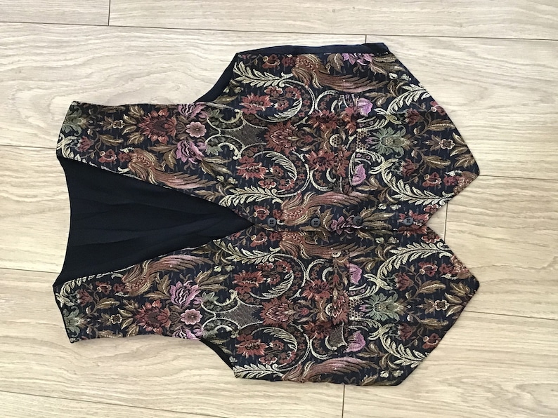 K&ouml;nnte beinhalten: Eine &auml;rmellose Weste mit dunkelblauer Grundfarbe und einem komplexen, mehrfarbigen Brokatmuster aus Blumen und V&ouml;geln in Rot-, Gold-, Gr&uuml;n- und Violettt&ouml;nen. Das Kleidungsst&uuml;ck verf&uuml;gt &uuml;ber einen V-Ausschnitt, eine Knopfleiste und zwei Vordertaschen.