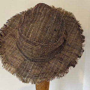 May include: A woven hat with a wide brim and a frayed edge. The hat is made of a textured, brown and tan material. The hat is displayed on a wooden stand.