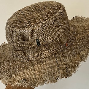 May include: A wide-brimmed hat made of woven, natural-toned fabric. The hat features a rounded crown and a frayed edge on the brim. A small black tag with white text is visible on the side of the hat.