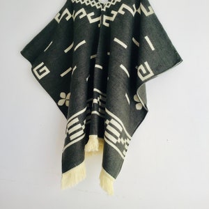 May include: A dark gray poncho with a geometric pattern in off-white. The poncho has a V-neck and fringe along the bottom edge. The design features rectangular and floral shapes. The poncho is hanging on a wooden hanger against a white background.