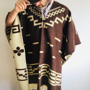 May include: A brown and cream-colored poncho with geometric patterns and fringe. The poncho has a dark brown base with cream-colored geometric designs. The edges are trimmed with cream-colored fringe. The poncho is worn over a white shirt.