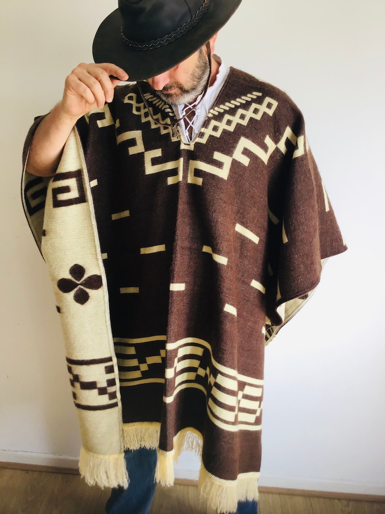 May include: A brown and cream-colored poncho with geometric patterns and fringe. The poncho has a dark brown base with cream-colored geometric designs. The edges are trimmed with cream-colored fringe. The poncho is worn over a white shirt.