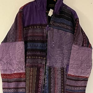 Può includere: Una giacca con cappuccio in patchwork viola con chiusura a zip. La giacca è realizzata con diversi tessuti con vari motivi e colori, tra cui viola, nero, rosso e bianco.