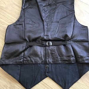 K&ouml;nnte beinhalten: Eine schwarze Weste mit einem glatten, satinartigen R&uuml;ckenteil, einem verstellbaren Riemen und einer silberfarbenen Metallschnalle. Der untere Teil der Weste hat eine matte schwarze Stoffoberfl&auml;che mit einem einzelnen Knopf am unteren Saum.