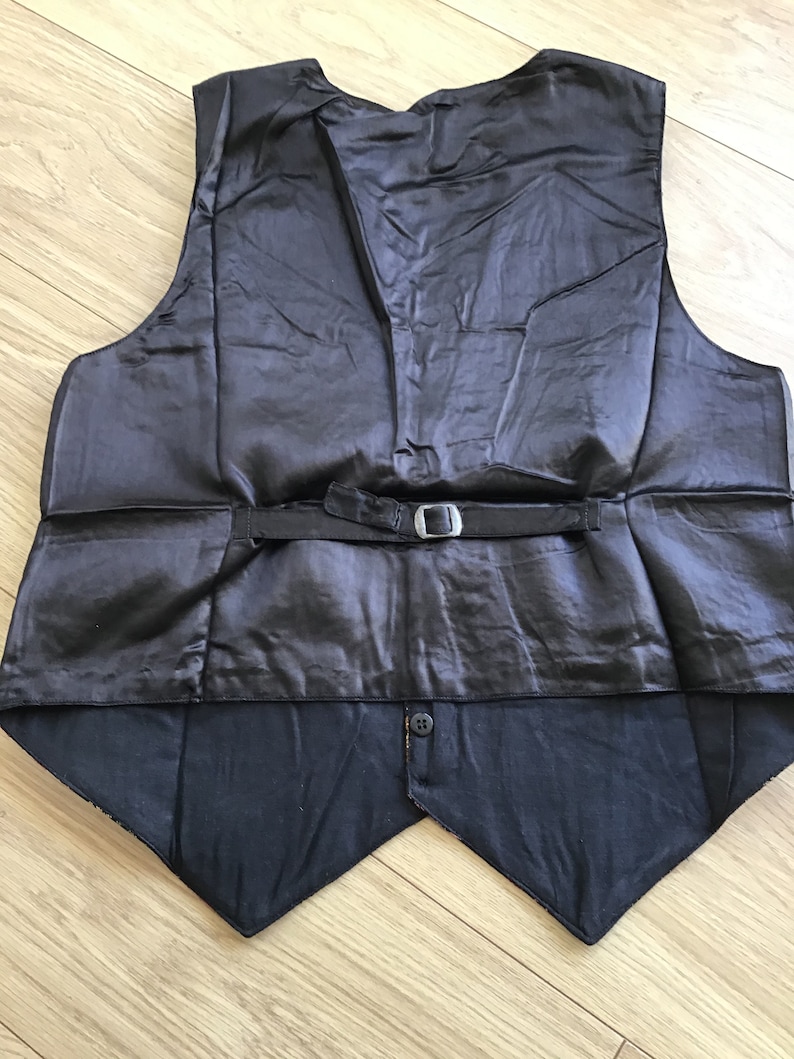 K&ouml;nnte beinhalten: Eine schwarze Weste mit einem glatten, satinartigen R&uuml;ckenteil, einem verstellbaren Riemen und einer silberfarbenen Metallschnalle. Der untere Teil der Weste hat eine matte schwarze Stoffoberfl&auml;che mit einem einzelnen Knopf am unteren Saum.