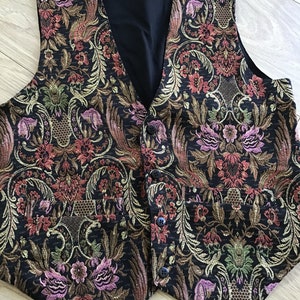 K&ouml;nnte beinhalten: Eine &auml;rmellose Weste mit schwarzer Basis und einem komplizierten, mehrfarbigen Blumen- und Paisley-Jacquardmuster in Rosa-, Gr&uuml;n- und Goldt&ouml;nen. Das Kleidungsst&uuml;ck hat einen V-Ausschnitt, vier dunkle Kn&ouml;pfe auf der Vorderseite und zwei Taschen.