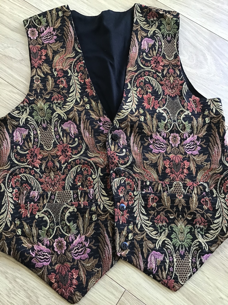 K&ouml;nnte beinhalten: Eine &auml;rmellose Weste mit schwarzer Basis und einem komplizierten, mehrfarbigen Blumen- und Paisley-Jacquardmuster in Rosa-, Gr&uuml;n- und Goldt&ouml;nen. Das Kleidungsst&uuml;ck hat einen V-Ausschnitt, vier dunkle Kn&ouml;pfe auf der Vorderseite und zwei Taschen.