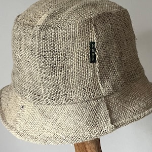 May include: A beige bucket hat made of a woven hemp fabric. The hat has a wide brim and a rounded crown. A small green tag with the word "HEMP" is sewn on the side. The hat is displayed on a wooden stand.