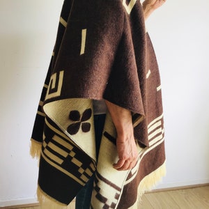 May include: A brown and cream-colored poncho with geometric patterns and fringe. The poncho features a repeating design of rectangles and squares in cream against a brown background. The fringe is a light cream color.