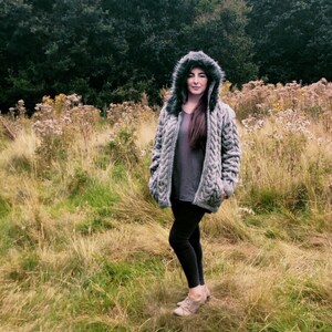 Puede incluir: Un cárdigan gris de punto de cable con capucha y ribete de piel sintética alrededor de la capucha. El cárdigan se lleva sobre una camiseta gris y leggings negros.