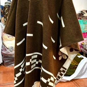 May include: A brown poncho with white geometric patterns and fringe trim. The poncho is displayed on a mannequin, showcasing its design and texture. The design includes rectangular and diagonal shapes.