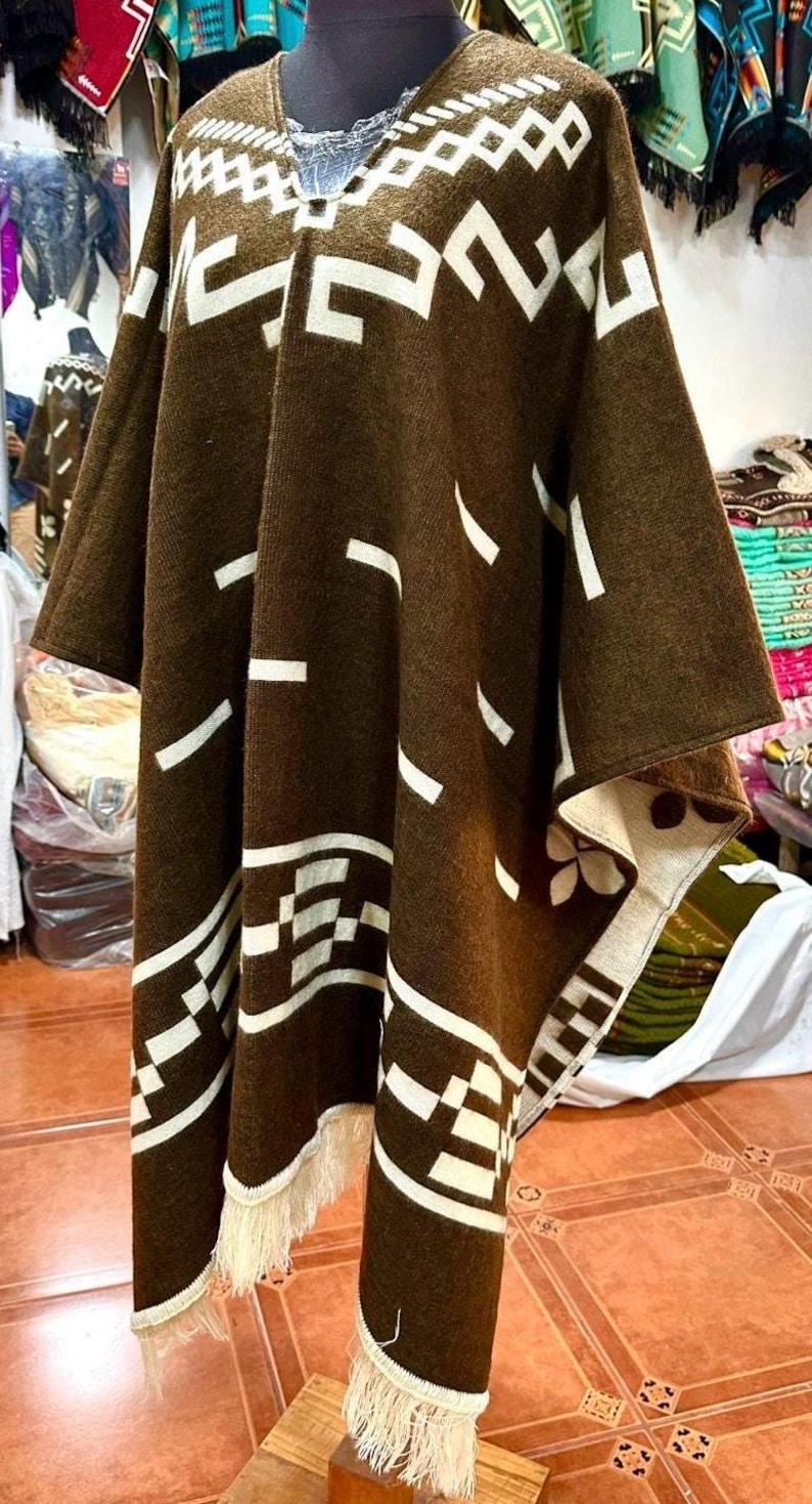 May include: A brown poncho with white geometric patterns and fringe trim. The poncho is displayed on a mannequin, showcasing its design and texture. The design includes rectangular and diagonal shapes.