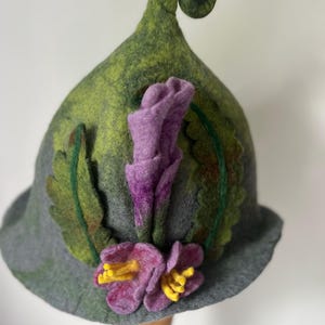May include: A whimsical felt hat in shades of green and gray, adorned with purple and yellow flowers. The hat features a curled, pointed top and is decorated with felt leaves. The overall design is reminiscent of a fairy or woodland theme.