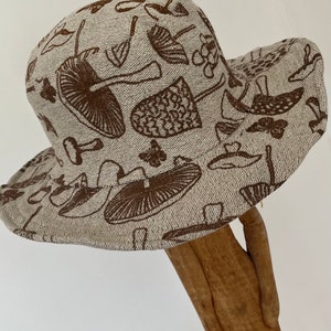 May include: A beige linen bucket hat with a brown mushroom print. The hat has a wide brim and a flat crown.