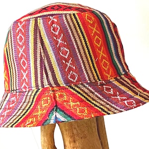 May include: A colorful bucket hat with a vibrant striped pattern in red, yellow, purple, and green. The hat features a repeating "xoxo" design and is displayed on a wooden stand. The brim is slightly angled.