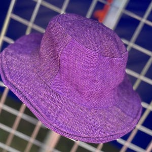 Puede incluir: Un sombrero morado vibrante con ala ancha. El sombrero está hecho de un material tejido, y la corona tiene un diseño ligeramente hundido. El sombrero está diseñado para dar sombra del sol.