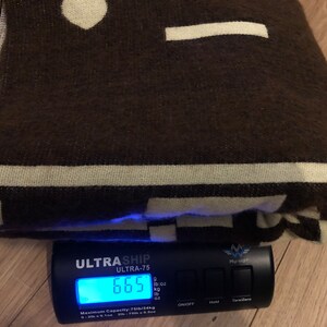 May include: A folded brown blanket with cream-colored geometric patterns and stripes. The blanket is on a scale that reads 6.65 lbs. The blanket has a soft, textured appearance, suggesting a cozy and warm textile.