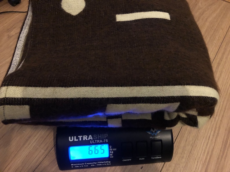 May include: A folded brown blanket with cream-colored geometric patterns and stripes. The blanket is on a scale that reads 6.65 lbs. The blanket has a soft, textured appearance, suggesting a cozy and warm textile.