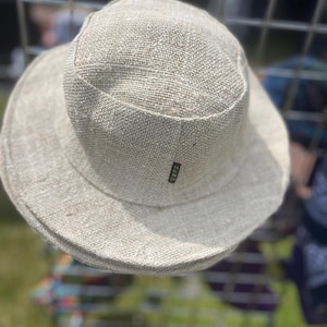 May include: A light beige bucket hat made of woven fabric. The hat has a wide brim and a rounded crown. A small black tag is sewn on the side of the hat. The hat is displayed against a blurred background.