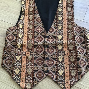 K&ouml;nnte beinhalten: Eine &auml;rmellose Weste mit einem komplexen geometrischen Muster in Braun-, Beige- und Schwarzt&ouml;nen. Das Kleidungsst&uuml;ck hat einen V-Ausschnitt, ein einfarbig schwarzes Innenfutter, vier dunkle Kn&ouml;pfe an der Vorderseite sowie zwei Vordertaschen.