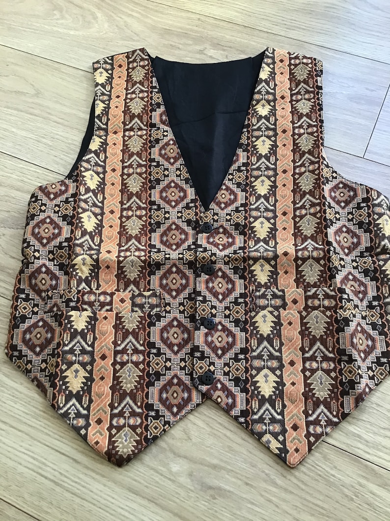 K&ouml;nnte beinhalten: Eine &auml;rmellose Weste mit einem komplexen geometrischen Muster in Braun-, Beige- und Schwarzt&ouml;nen. Das Kleidungsst&uuml;ck hat einen V-Ausschnitt, ein einfarbig schwarzes Innenfutter, vier dunkle Kn&ouml;pfe an der Vorderseite sowie zwei Vordertaschen.
