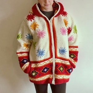 May include: A white knitted cardigan with a colourful floral pattern and a red, orange and green geometric pattern at the bottom. The cardigan has a hood and a zip closure.