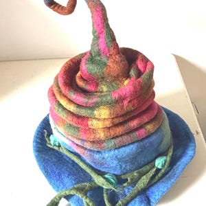 May include: A felted hat with a wide blue brim and a tall, spiraling, pointed crown. The crown features a variegated pattern of pink, orange, yellow, and green wool fibers. A thin, green, vine-like felted cord with small buds wraps around the base.
