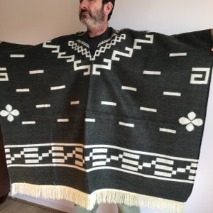 May include: Dark gray poncho with white geometric patterns and fringe. The poncho features a repeating design of rectangles, squares, and floral shapes. The fringe is a light cream color. This poncho is a stylish outerwear garment.