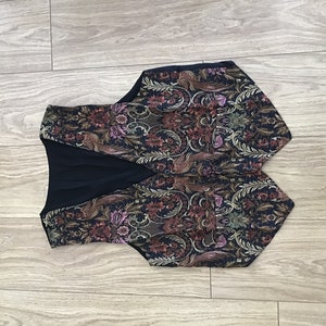 K&ouml;nnte beinhalten: Eine &auml;rmellose Weste mit dunklem Stoffgrund und einem komplexen, mehrfarbigen Blumen- und Paisley-Gobelinmuster. Das Kleidungsst&uuml;ck verf&uuml;gt &uuml;ber einen V-Ausschnitt, zwei kleine Vordertaschen und ein einfarbig schwarzes Innenfutter aus Stoff.