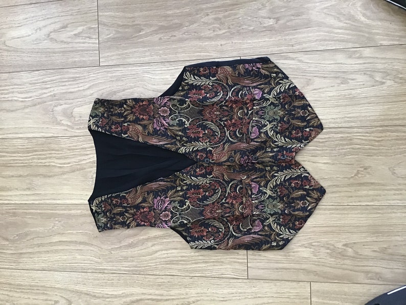 K&ouml;nnte beinhalten: Eine &auml;rmellose Weste mit dunklem Stoffgrund und einem komplexen, mehrfarbigen Blumen- und Paisley-Gobelinmuster. Das Kleidungsst&uuml;ck verf&uuml;gt &uuml;ber einen V-Ausschnitt, zwei kleine Vordertaschen und ein einfarbig schwarzes Innenfutter aus Stoff.