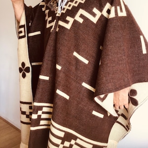May include: A brown and cream-colored poncho with geometric patterns and fringe. The poncho features a repeating design of squares and lines in cream against a brown background. The edges are trimmed with cream-colored fringe.