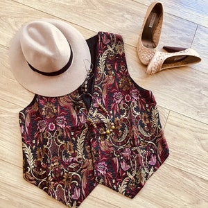 K&ouml;nnte beinhalten: Ein beiger Fedora-Hut mit schwarzem Band, eine &auml;rmellose Weste mit einem floralen Gobelinmuster in Dunkelrot, Gold und Schwarz, eine Perlenkette mit dunklen und metallischen Perlen sowie ein Paar beige geflochtene Pumps mit braunen Abs&auml;tzen.