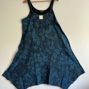 Puede incluir: Un vestido de peto de mezclilla negro con estampado floral azul. El vestido tiene tirantes ajustables y cierre de botón en los hombros.