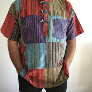 Puede incluir: Una camisa de manga corta de rayas multicolores con cuello mandarín y cierres de botones de madera. La camisa está hecha de un mosaico de rayas de diferentes colores.