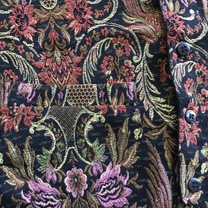 K&ouml;nnte beinhalten: Eine schwarze Weste mit einem gewebten floralen Tapisseriemuster in Rosa, Rot, Gold und Gr&uuml;n. Das Kleidungsst&uuml;ck verf&uuml;gt &uuml;ber eine Vordertasche und eine vertikale Reihe von vier kleinen, runden, schwarzen Kn&ouml;pfen an der rechten Stoffkante.
