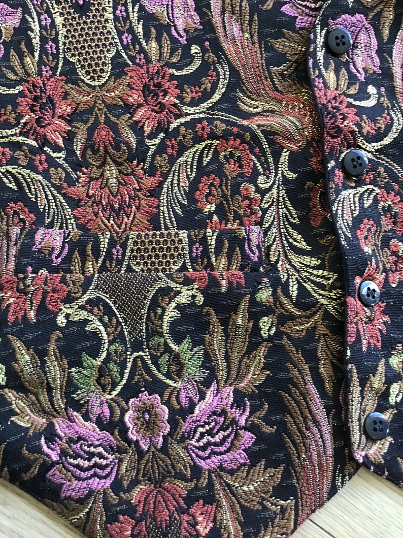 K&ouml;nnte beinhalten: Eine schwarze Weste mit einem gewebten floralen Tapisseriemuster in Rosa, Rot, Gold und Gr&uuml;n. Das Kleidungsst&uuml;ck verf&uuml;gt &uuml;ber eine Vordertasche und eine vertikale Reihe von vier kleinen, runden, schwarzen Kn&ouml;pfen an der rechten Stoffkante.